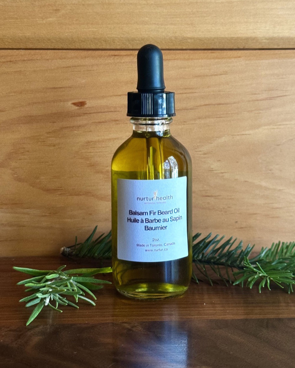 Two-ounce bottle of Balsam Fir Beard Oil on a wood table, fir and rosemary sprigs alongside