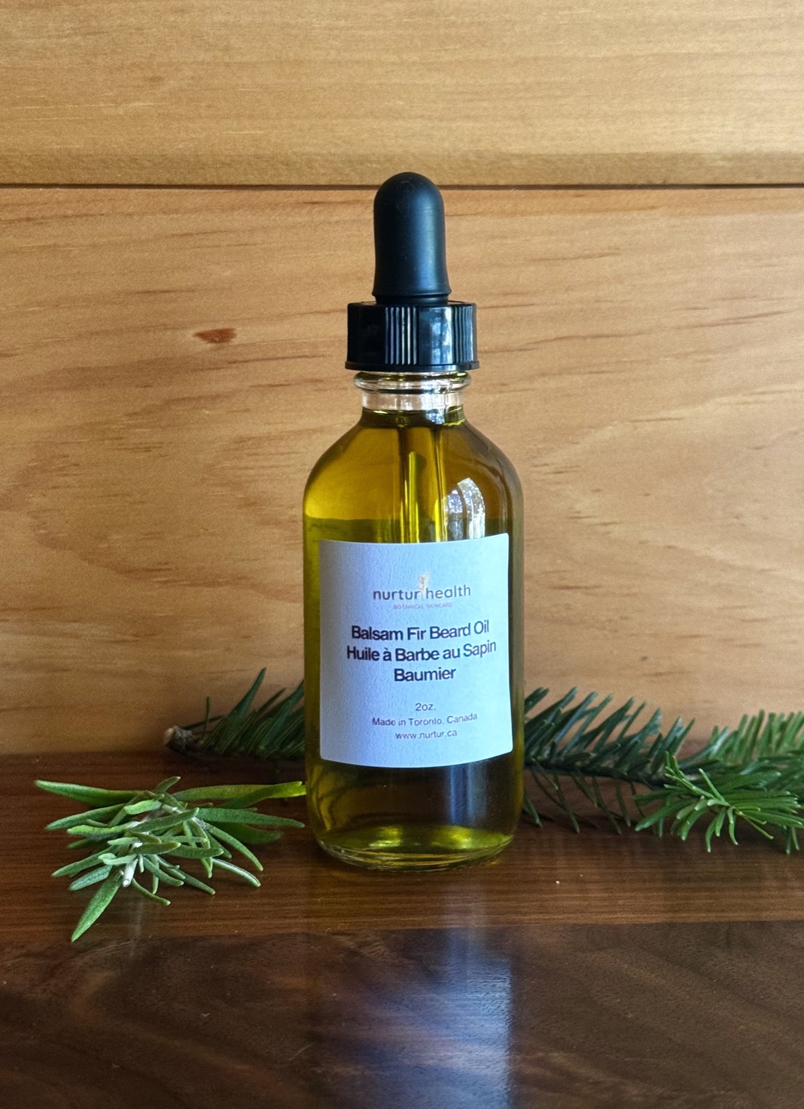 Two-ounce bottle of Balsam Fir Beard Oil on a wood table, fir and rosemary sprigs alongside