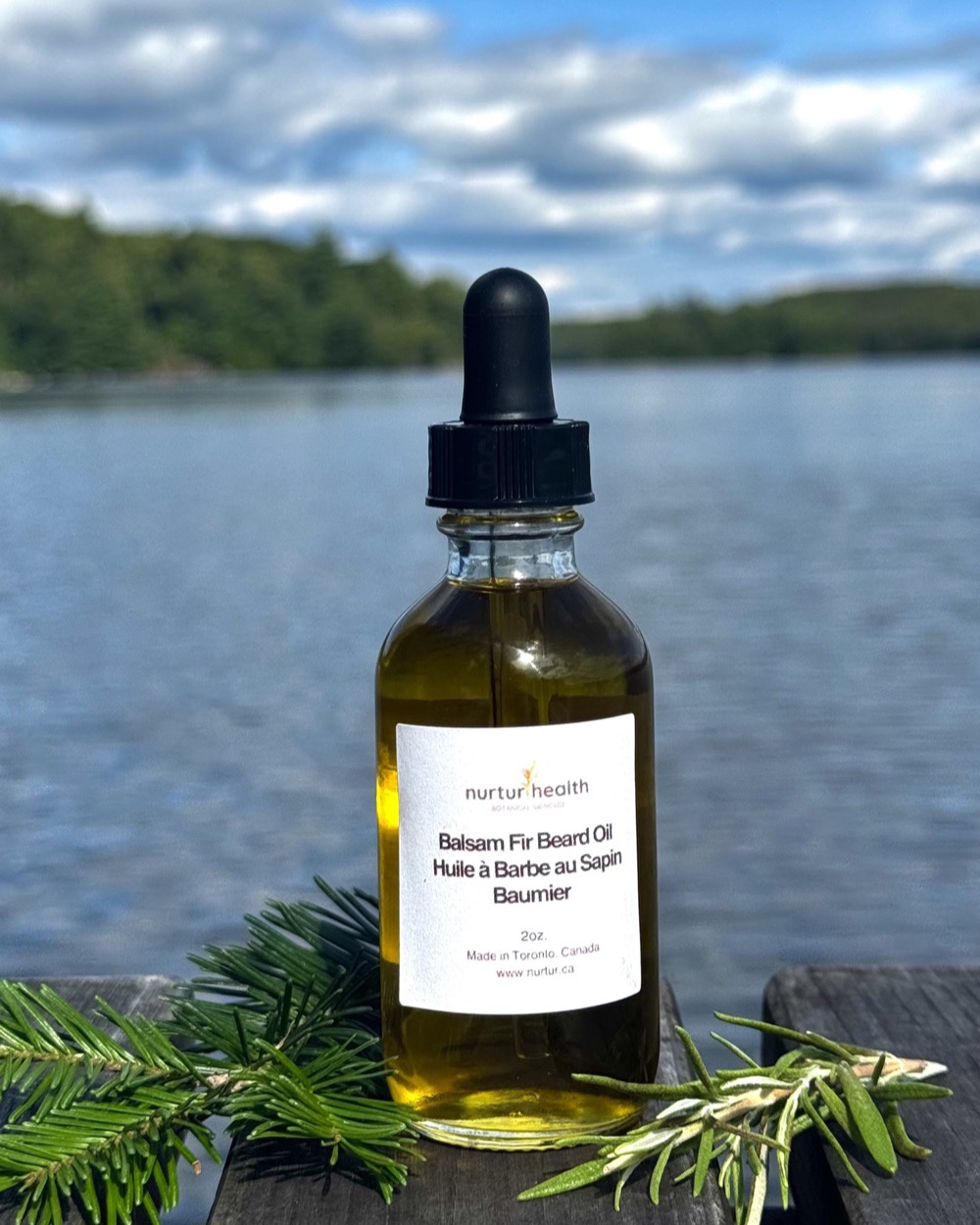 The bottle on a dock above a lake, fir and rosemary sprigs alongside