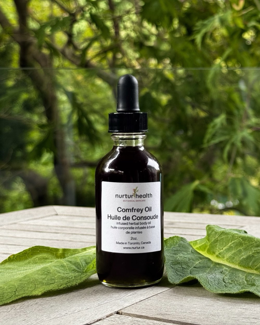 Two-ounce amber dropper bottle of Comfrey Oil on a weathered wood table, fresh comfrey leaves alongside